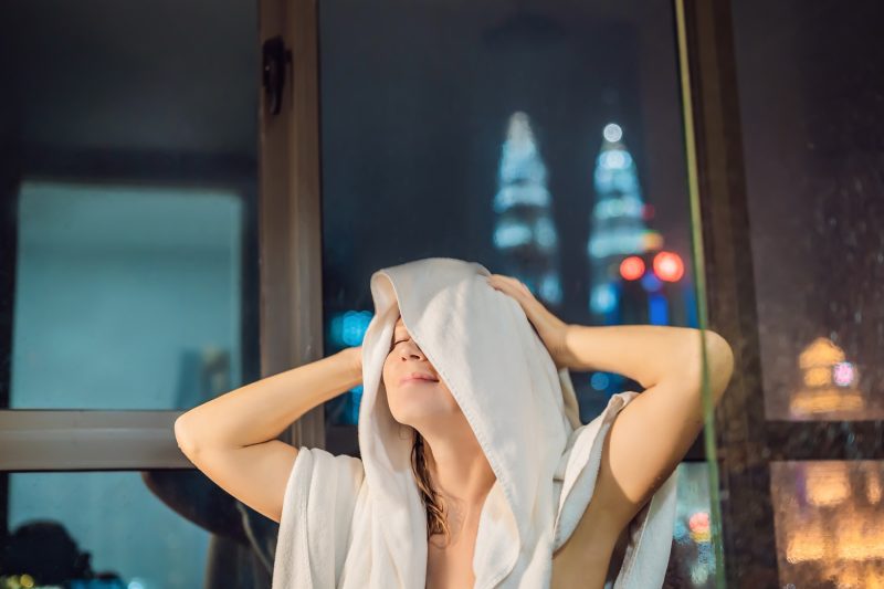 Beautiful woman wipes her hair after a shower on the background of a window with a panoramic view of the night city.